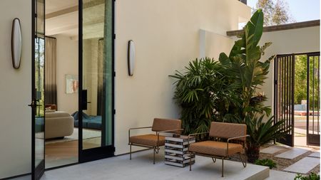 An outdoor patio off the side of a large, contemporary, off-white home with black trim. There are two burnt orange outdoor chairs, a side table, and sconce lights on the wall framing the glass door.