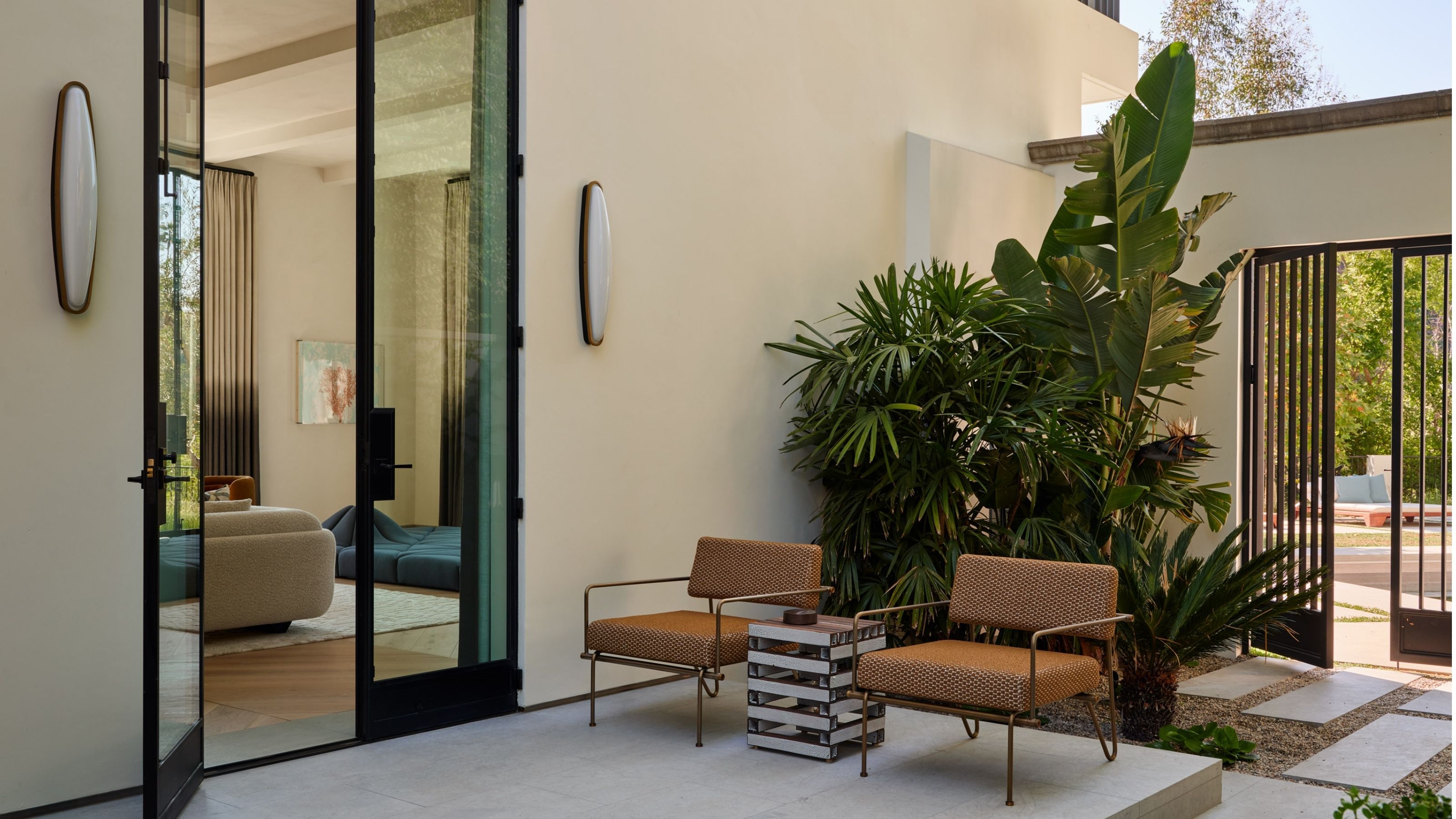 An outdoor patio off the side of a large, contemporary, off-white home with black trim. There are two burnt orange outdoor chairs, a side table, and sconce lights on the wall framing the glass door.