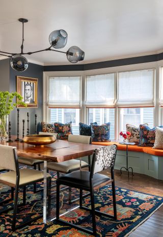 Image of a colorful, and patterned living room. There is a multicolored rug under the dining table, cow print chairs, and a window seat with a bright orange cushion.