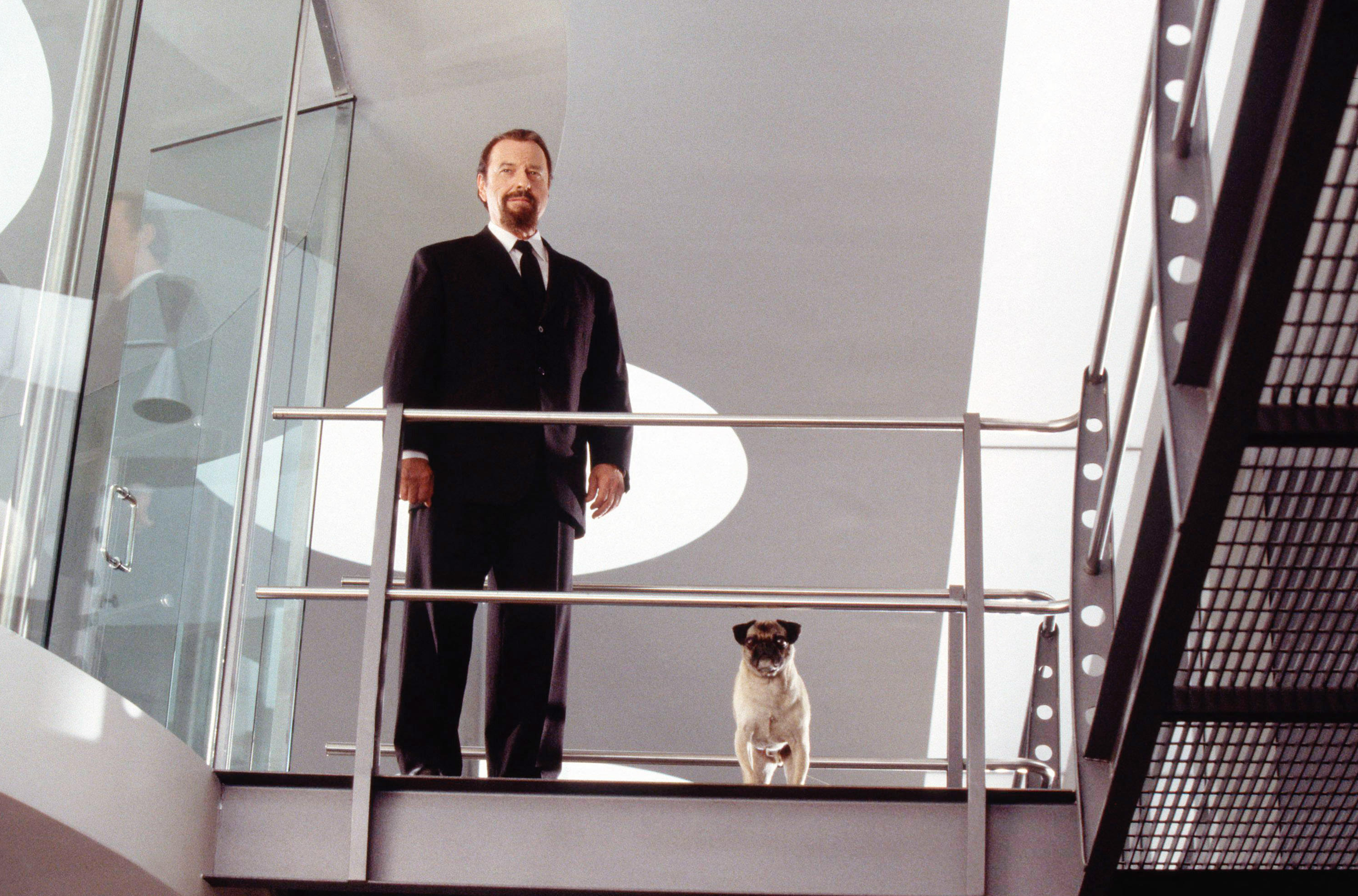A man in a black suit stands on a high walkway in a futuristic headquarters with Frank the Pug at his side, both looking down over the railing.