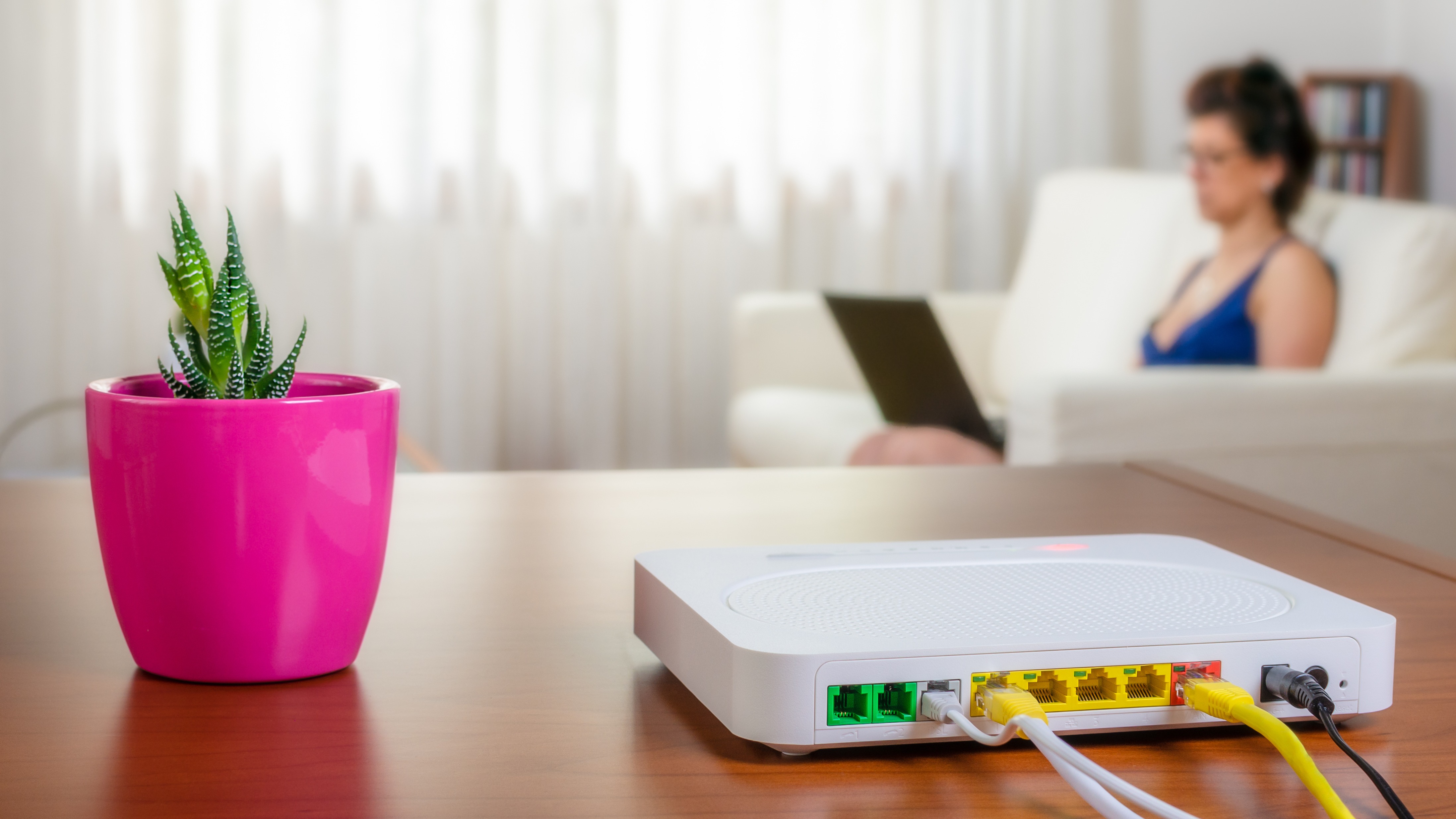 Modem router on a table in a living room. A woman using a laptop while sitting on the sofa is in background. Selective focus.