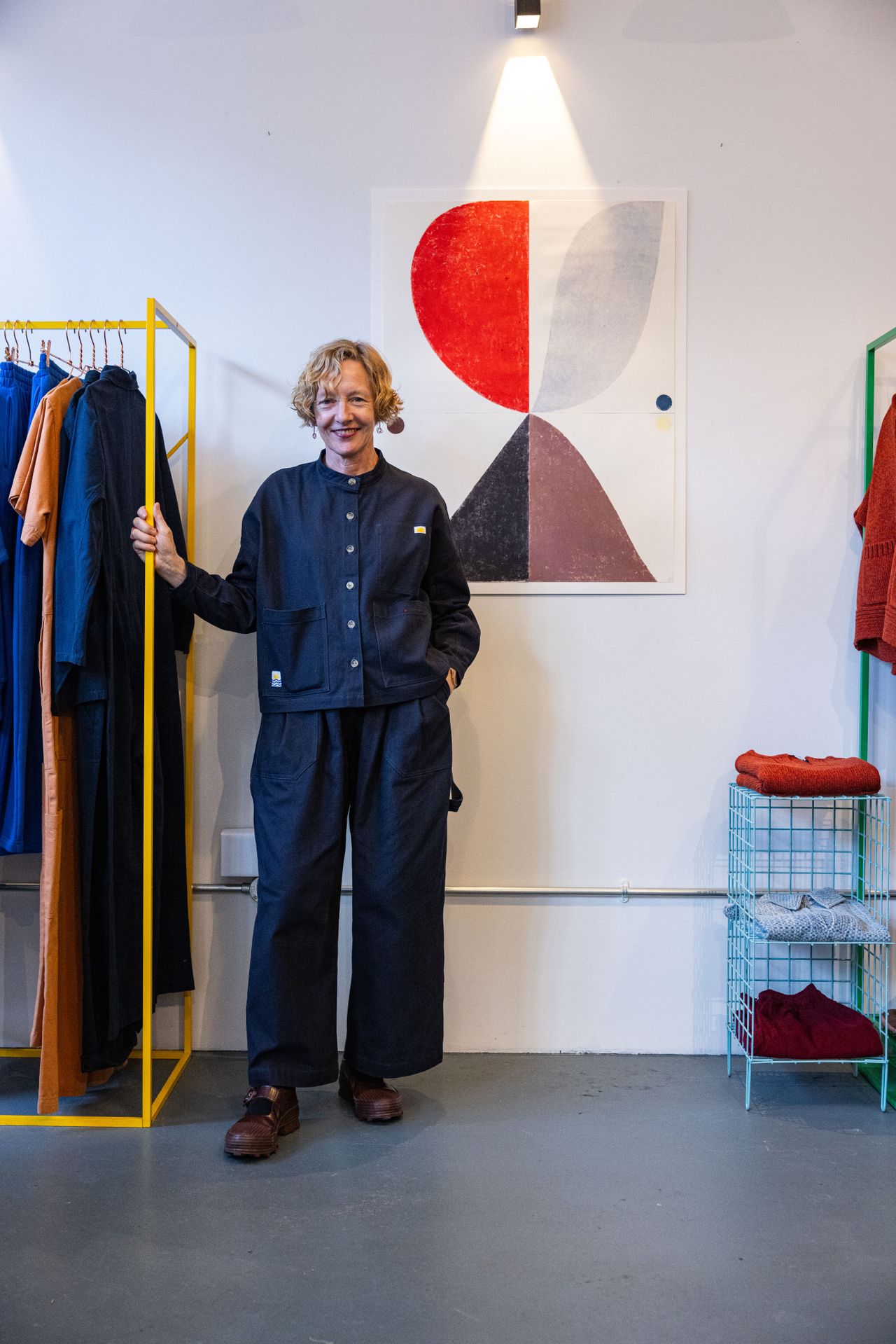 A clothing and design showroom hosts an exhibition of colorful, abstract prints now adorning its plain walls, immersed in colorful decor, and hosts the woman artist who designed them dressed in a double denim shirt and trousers outfit.