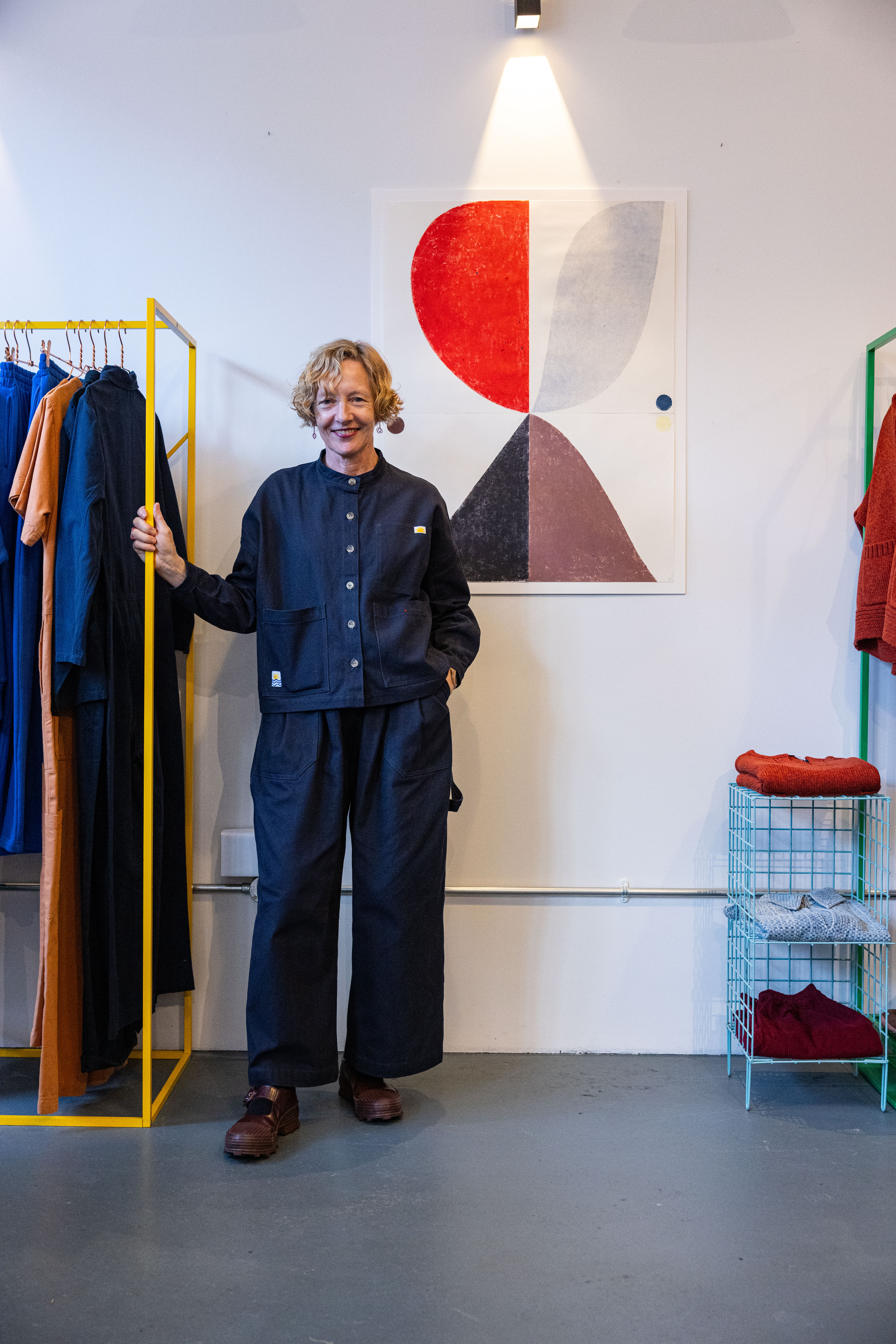 A clothing and design showroom hosts an exhibition of colorful, abstract prints now adorning its plain walls, immersed in colorful decor, and hosts the woman artist who designed them dressed in a double denim shirt and trousers outfit.