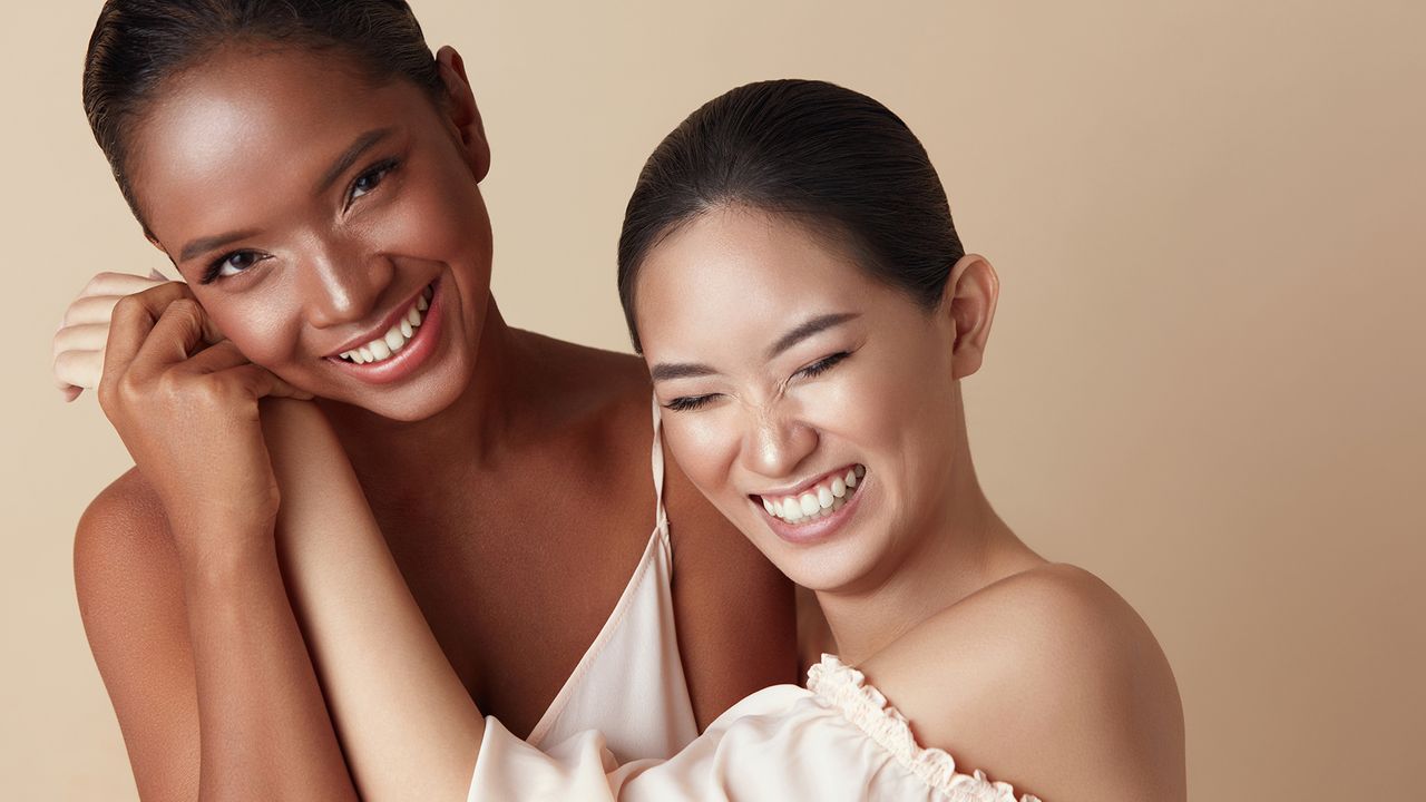 Diversity. Beauty Models Portrait. Cheerful Asian And Mixed Race Women Bonding Hands And Laughing. Different Ethnicity Female With Natural Makeup Standing Together Against Beige Background. - stock photo