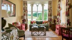 Warm bright living room with pale yellow walls, and red and green accents. There are multiple mismatched sofas and seats, with ornate metal coffee table and patterned curtains, cushions and rugs