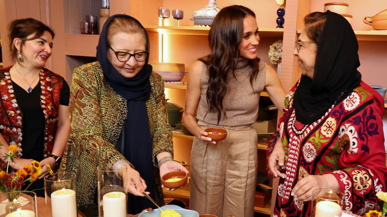 Meghan Markle smiling and putting her arm around a woman wearing a black head dress as two other women watch on and serve themselves dessert from a candle lit table