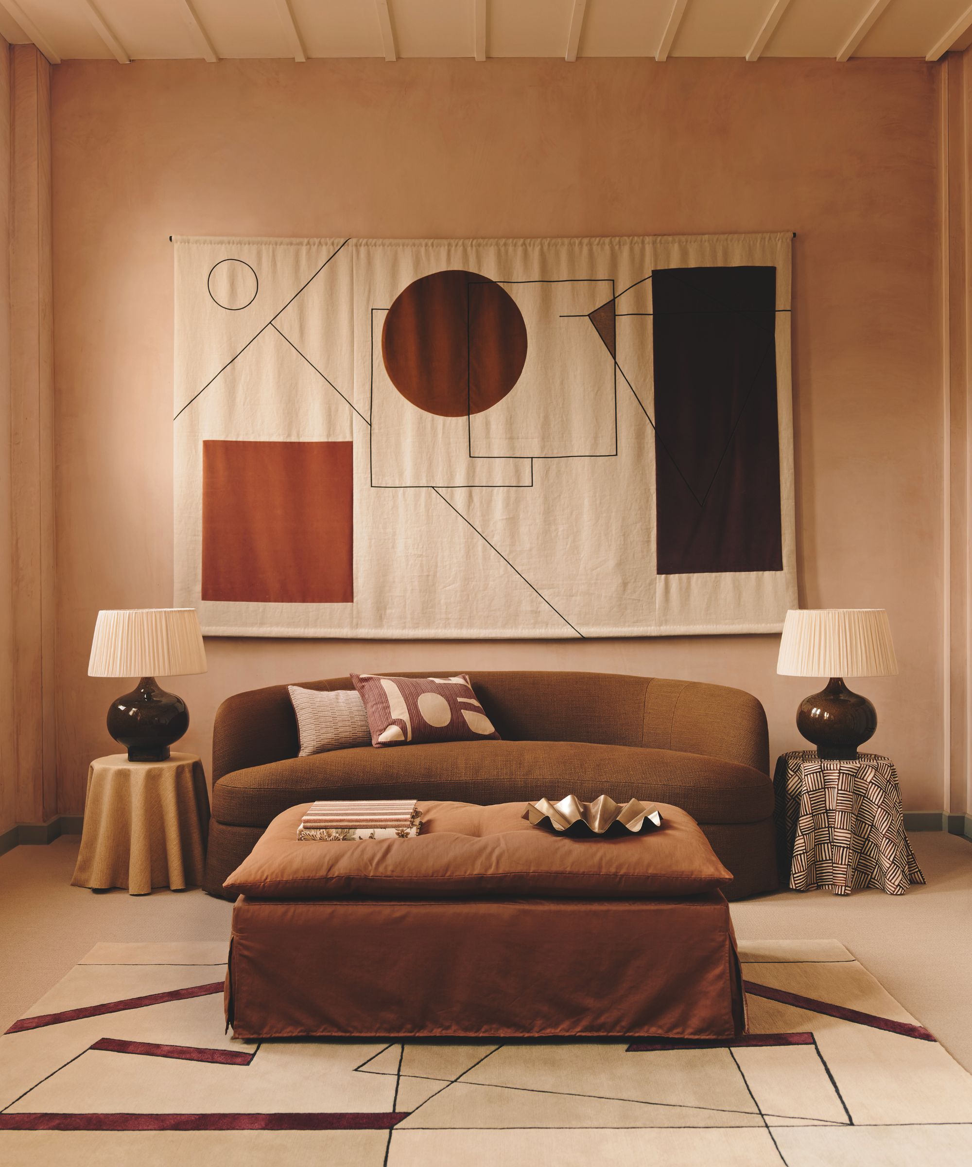 A warm, terracotta-toned living room featuring a curved chocolate-brown sofa and a large, abstract geometric wall hanging. A rust-colored ottoman sits in the center on a patterned rug, flanked by two table lamps with pleated shades.