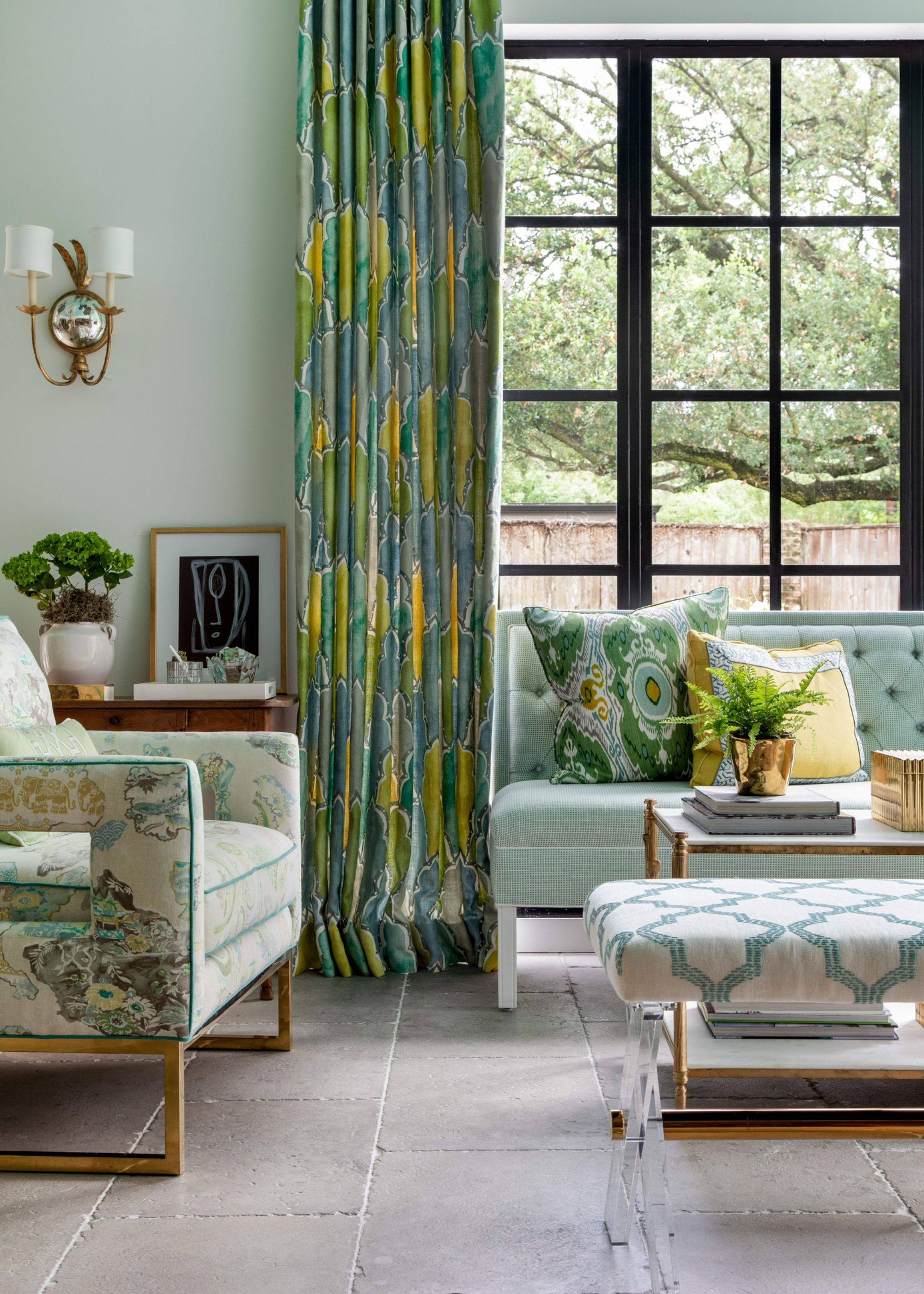 A light and airy living room with stone flooring, French doors with a black metal frame, light green walls, a mint green sofa and a floral green armchair, and green and yellow patterned curtains.