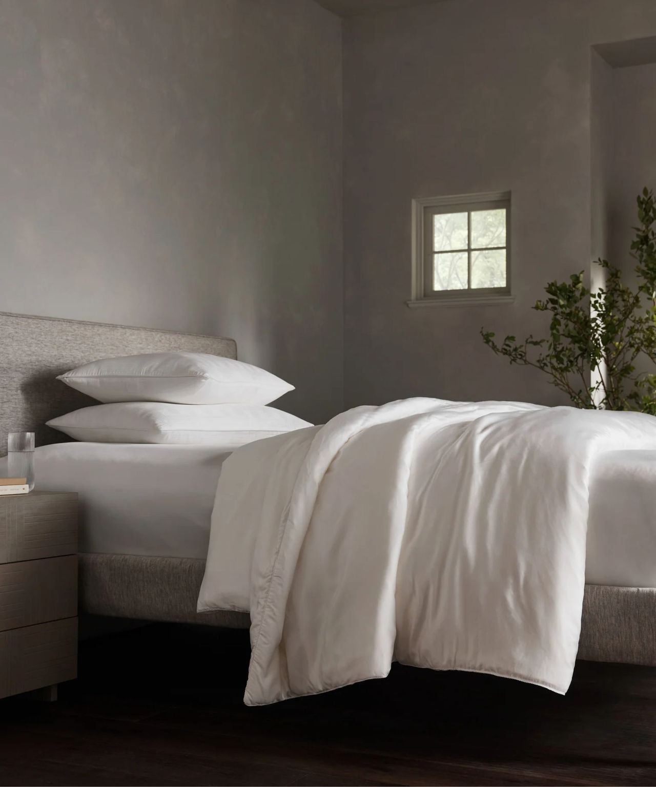 The Cozy Earth Silk Comforter pictured on a white mattress with white pillows, on a brown upholstered bed. To the right of the bed, in the foreground, half a chest of drawers is seen with a glass of water on top. In the background is a small window and a large tree. The walls are grey.