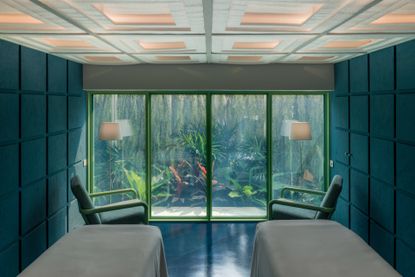 Interior of a treatment room at Infinity Wellbeing, Bangkok with dark teal square wall panelling, treatment tables, chairs and view of outside greenery
