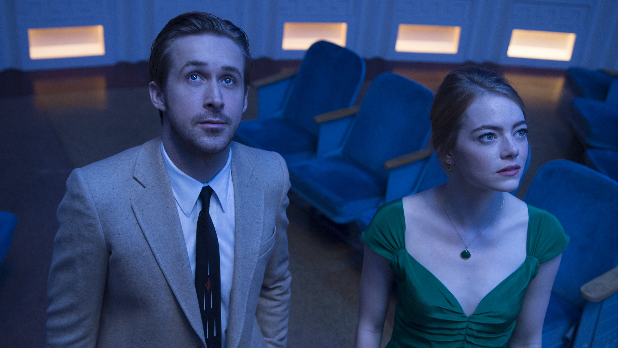 La La Land still. Mia and Sebastian stand in front of a row of blue chairs. Sebastian wears a cream suit with a black tie, and Mia wears a green dress,