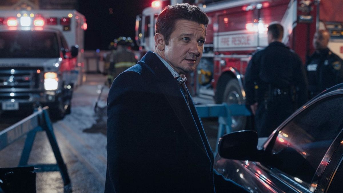 Jeremy Renner as Mike turning to his right and standing in front of an ambulance and fire truck.