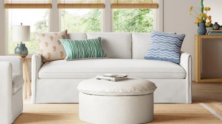 White sofa in living room with round upholstered ottoman and green and blue throw cushions, rattan venetian blinds