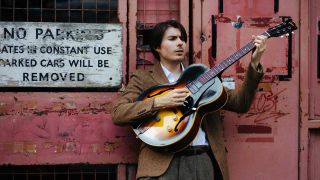 Connor Selby leaning against a entry to a decrepit building holding his guitar