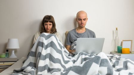A man and woman lying in bed together, with the man on his laptop and his wife looking angry