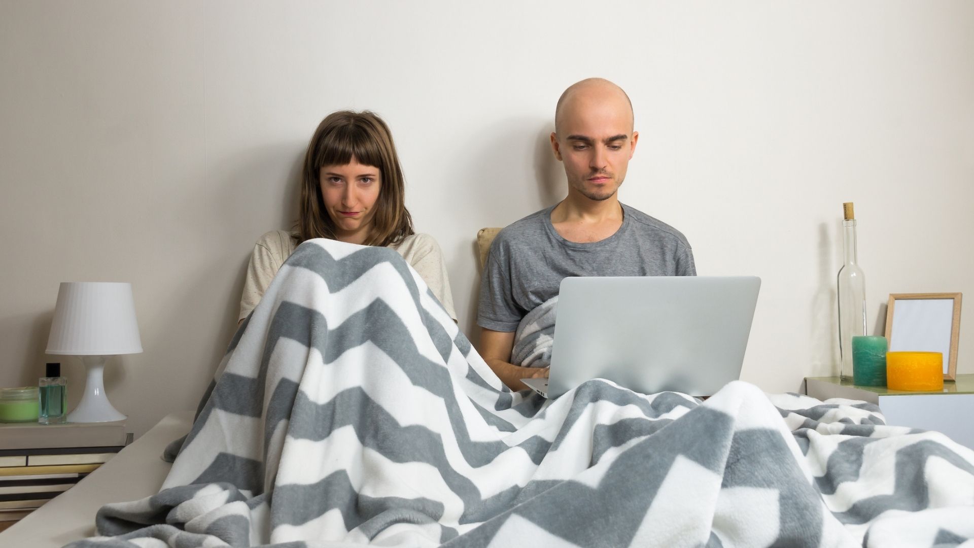 A man and woman lying in bed together, with the man on his laptop and his wife looking angry