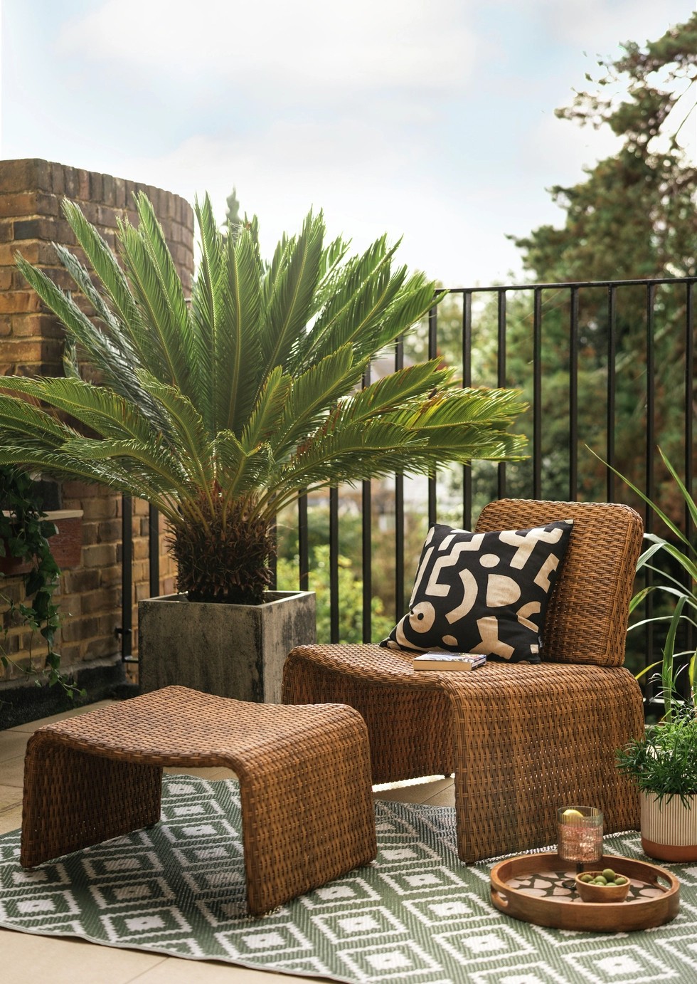 Image of an outdoor balcony with a large plant in the corner and a green and white diamond pattern rug. There is a brown basketweave chair and matching ottoman