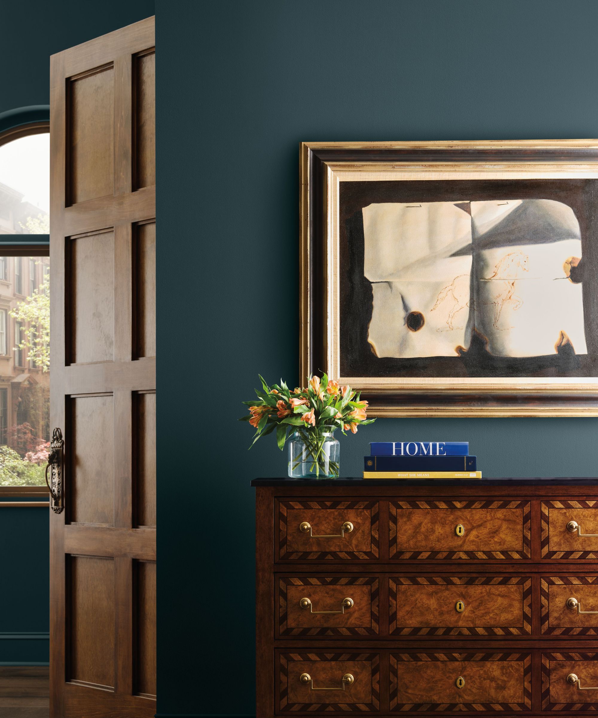 Dark navy painted walls with chest of drawers, abstract art on wall, stack of books and vase of flowers