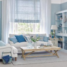 Calm and decluttered living room decorated in a relaxed coastal style, sofa and armchair with cushions in pale and mid blue, storage unit on the wall and pale blue wallpaper.
