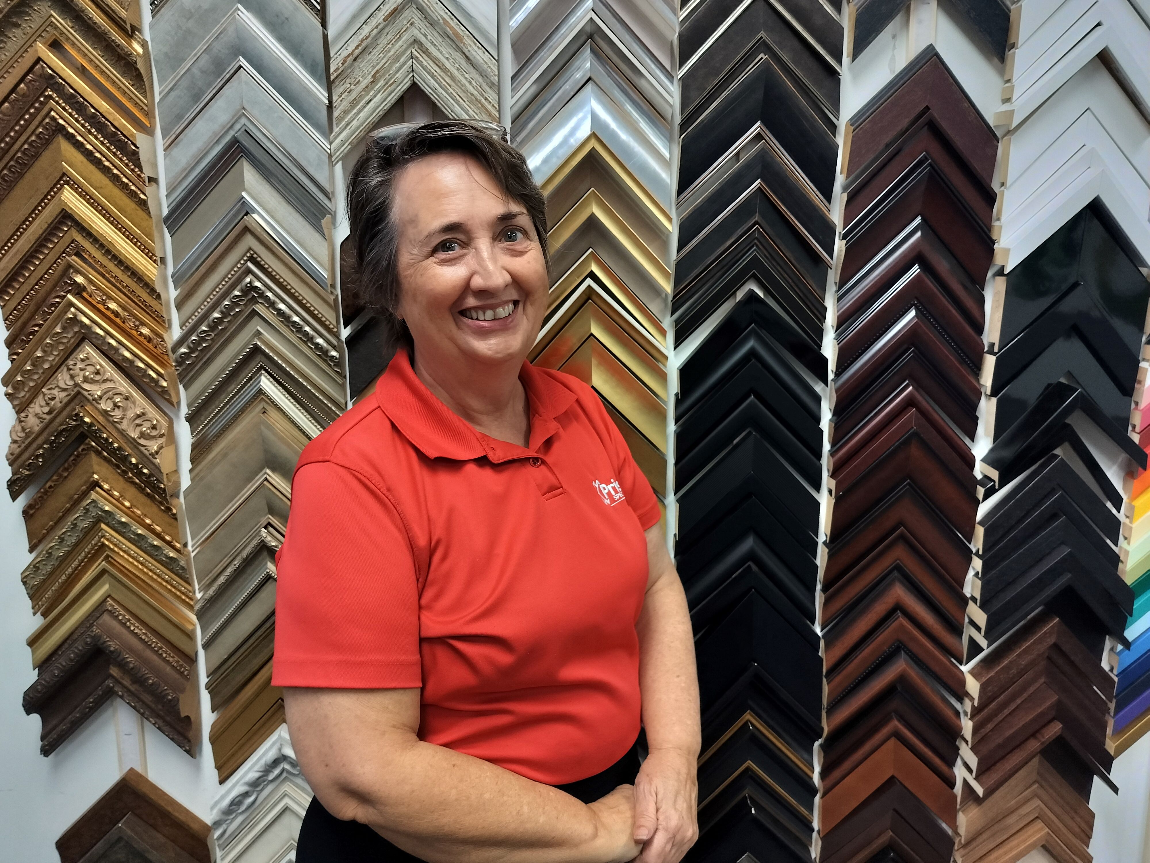 Smiling woman with short gray hair wearing red polo shirt, smiling in front of trim samples on wall is Jennifer Brashear of Prism Specialties