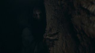 A creepy hooded figure hides in the shadows. They are peeking out from behind a tree, their long claw-like fingers holding onto the tree.