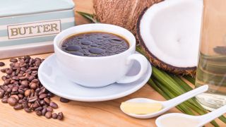 A cup of coffee with a teaspoon of butter, coffee beans, and a coconut around the edges