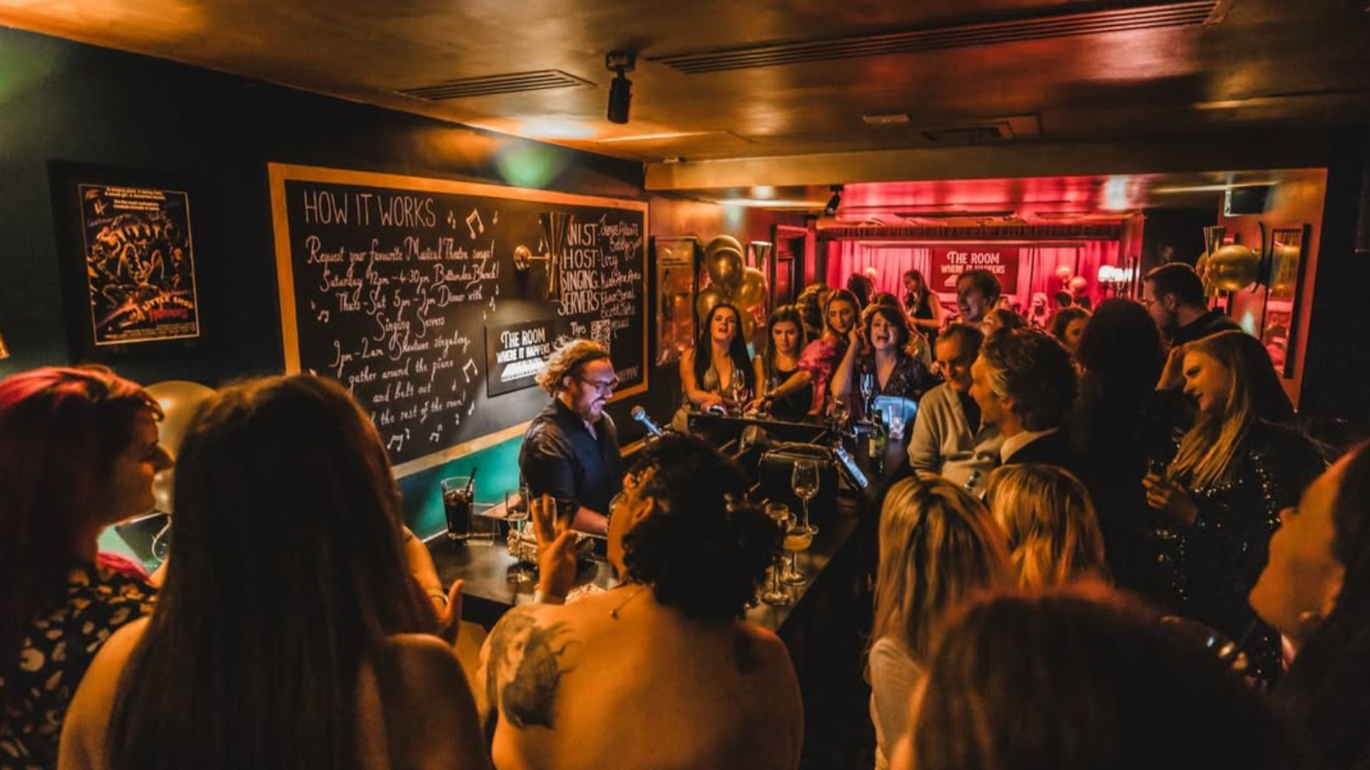 an image of the Room Where it Happens which is filled with people standing round a piano