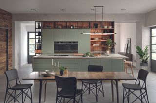 An open plan white kitchen with pale green cabinetry and island, a long wooden dining table and open wooden shelving.