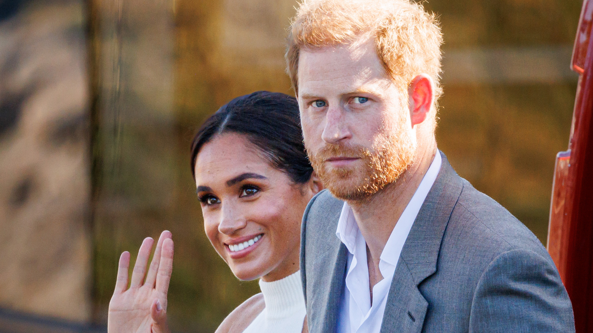 Meghan Markle wears a white halter top and waves while Prince Harry, who has red hair, wears a gray suit jacket and white shirt