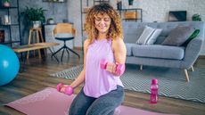 A woman kneels on an exercise mat holding light dumbbells in her hands in a modern-looking living room. There's a couch, desk chair and shelving behind her.
