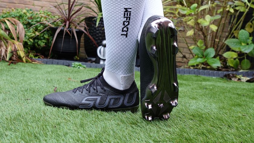 A person in long white socks is wearing black football boots and is showing the silver underside of one of the boots while on green artificial grass.