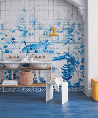 Bathroom with blue tiled floor, white double hand basin against white tiles with blue garden scene with birds.