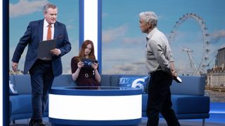 Hugh Bonneville in a suit as Douglas gets up from a sofa where Karen Gillan sits in a maroon dress as Madeline and Ben Miles in a check shirt and dark trousers as Toby walks towards Douglas in Douglas is Cancelled