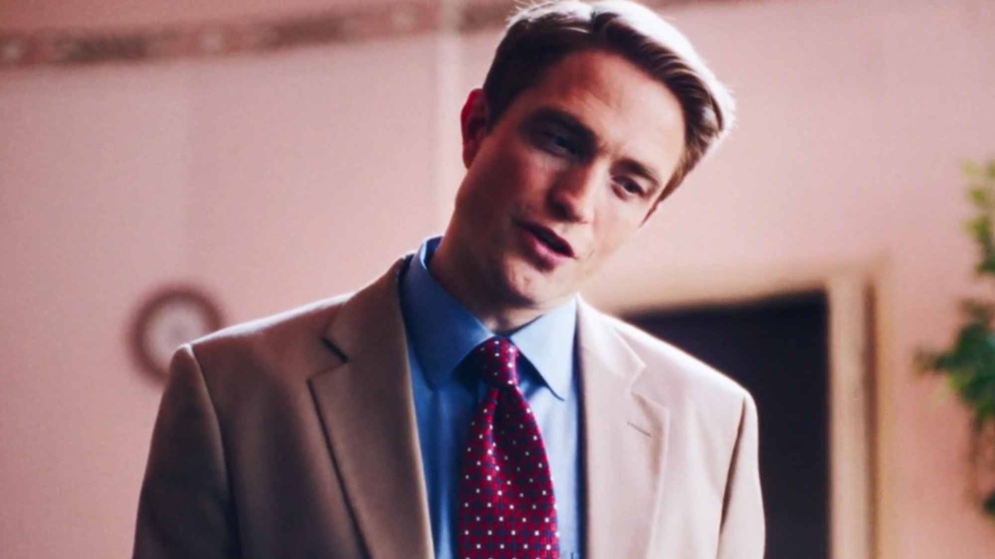 Robert Pattinson in a brown suit, blue shirt and red tie in Primetime. 