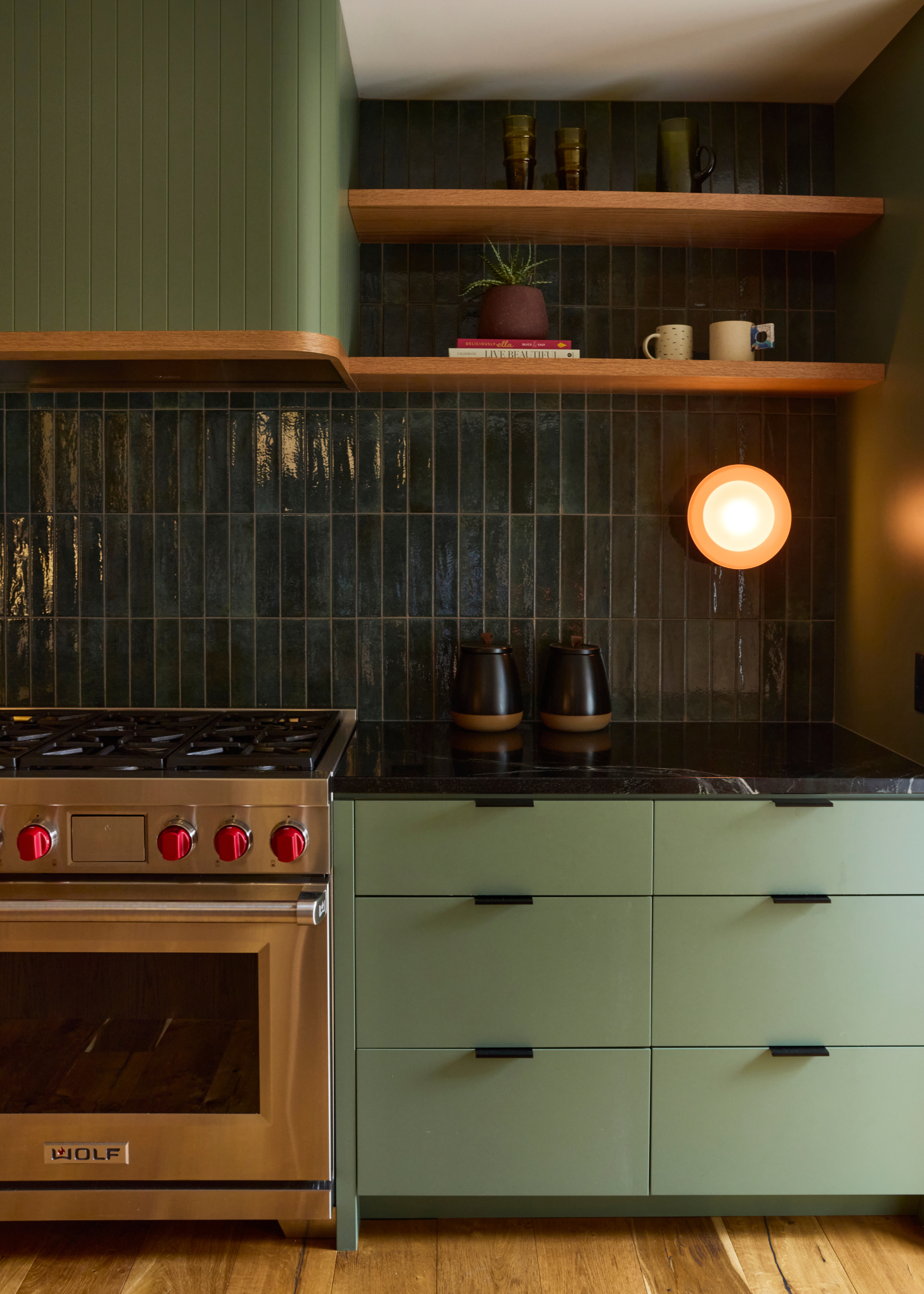 A kitchen with a green tile backsplash, wood floating shelves, a round sconce, a pair of black vessels, green drawers, and a steel stove and oven with red knobs below a tiled extractor hood