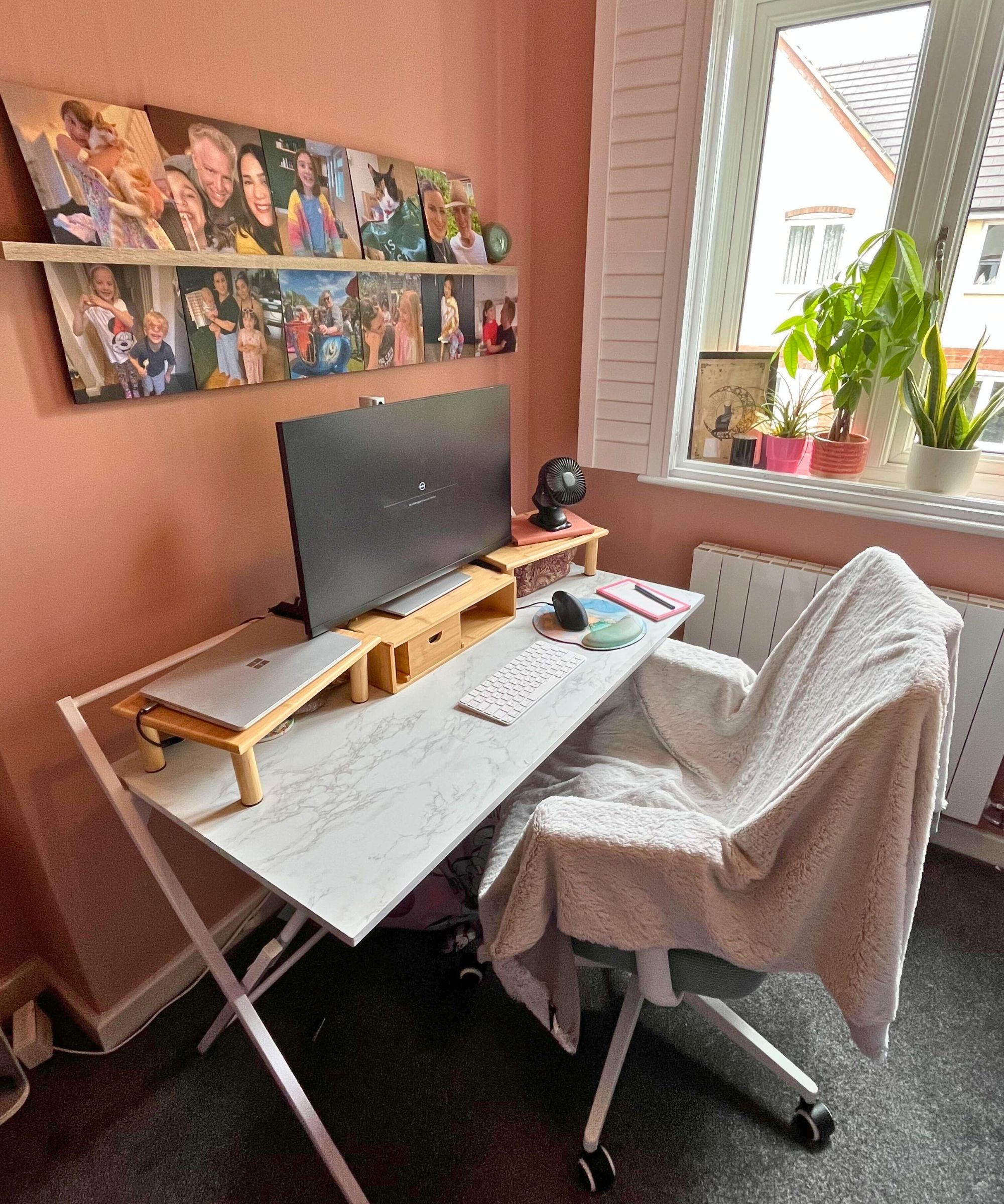 Terracotta colored painted walls in small home office with white marble effect desk, bamboo monitor riser and family pictures on the wall behind the monitor