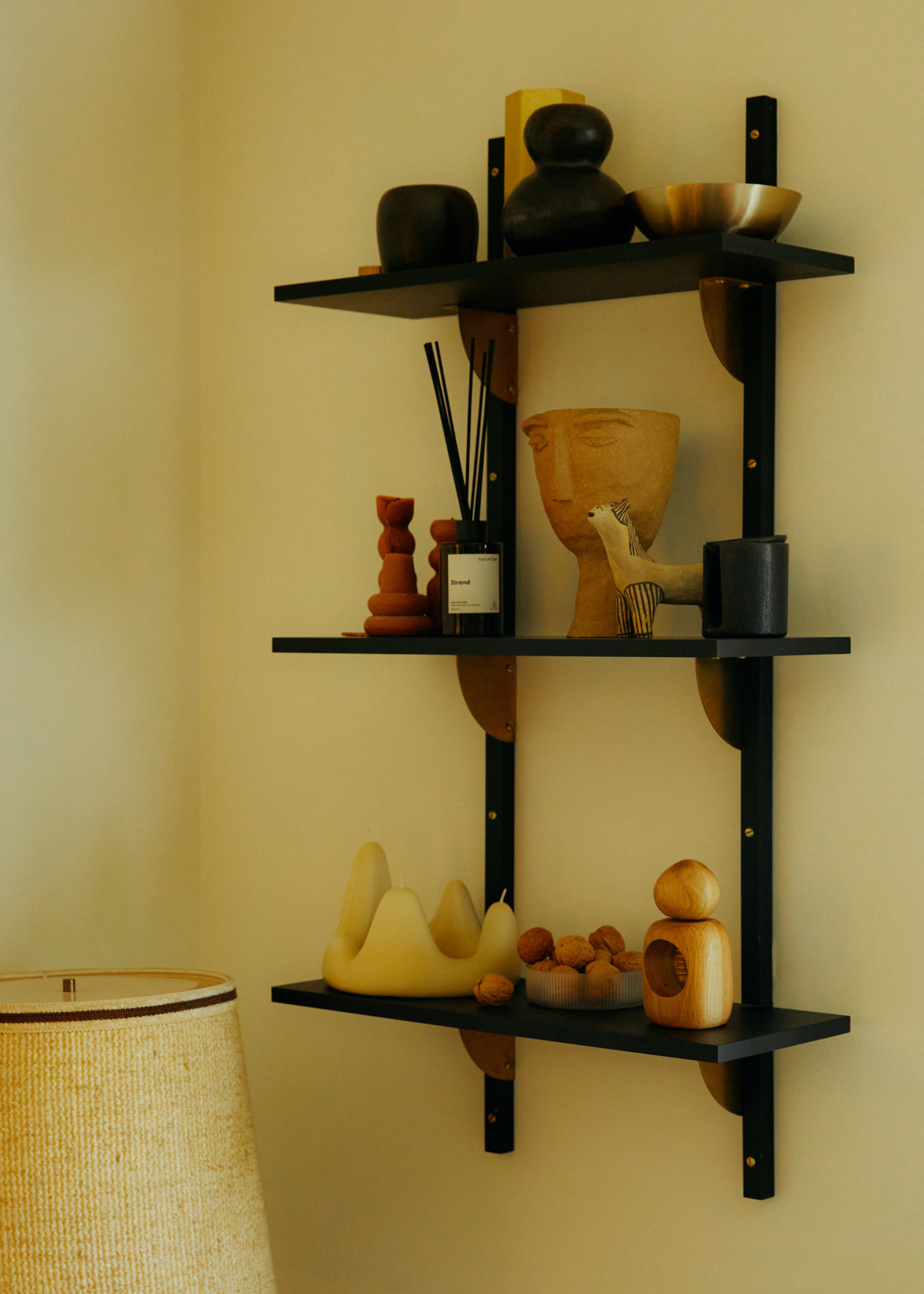 A hanging shelf by a lamp with three tiers featuring ceramics, candles, a brass bowl, a reed diffuser, and books