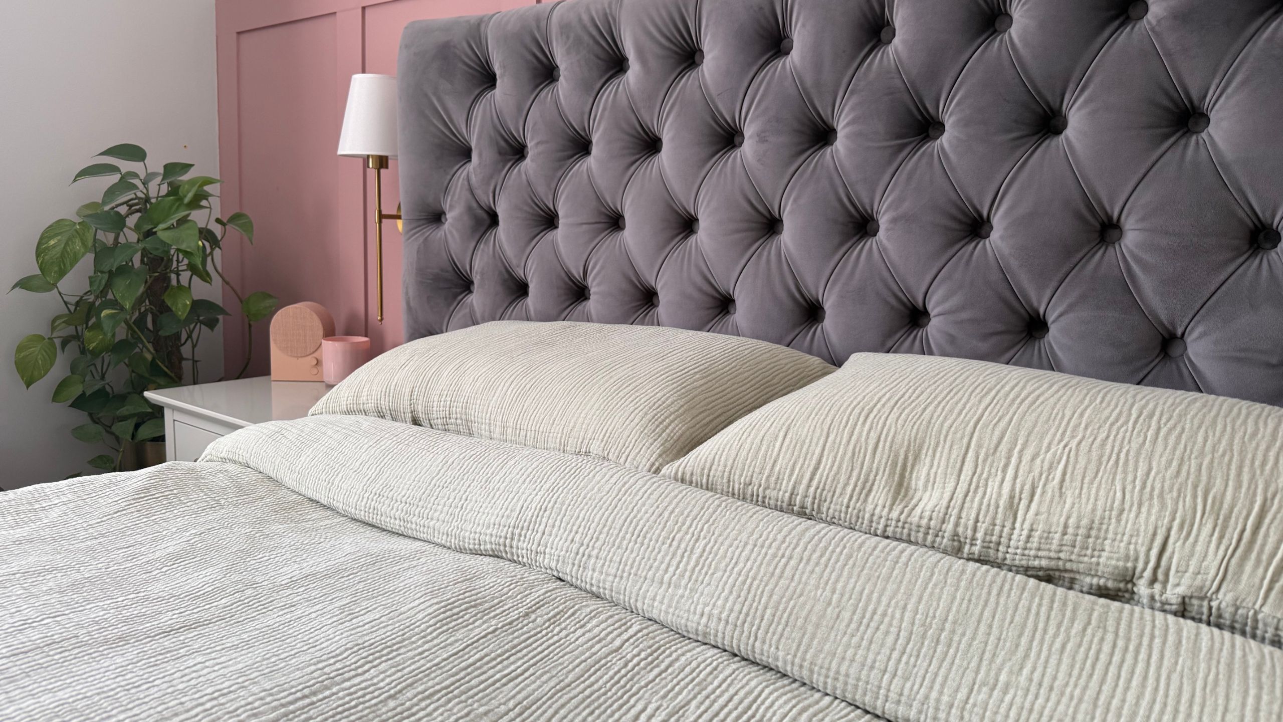 A close up of light green muslin bedding and two pillows on a bed with a dark blue-gray headboard and pink-painted wall-paneling behind