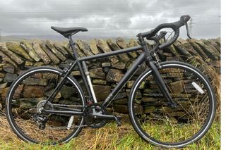 The black Brand X Road Bike has a frame pump secured between the top tube and bottom bracket photographed in front of a stone wall with grass under the wheels and grey sky above