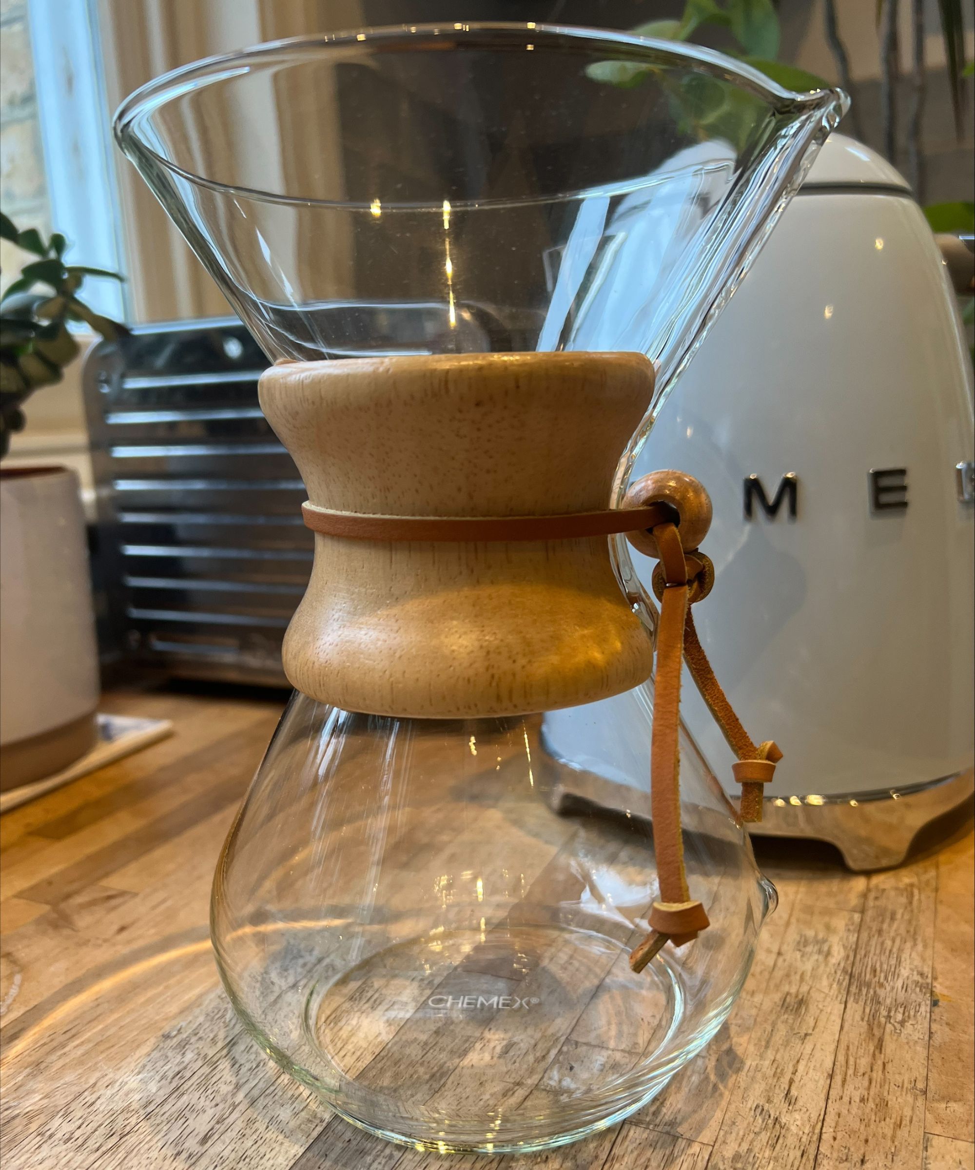 The CHEMEX coffee maker on a wooden countertop. Behind is a white pot with a small houseplant, a silver Dualit toaster, a baby blue Smeg kettle, larger houseplants, and a baby blue backsplash. To the left, a window surrounded by yellow brick is seen.