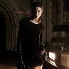 Model wearing a black AllSaints dress and gloves at night in front of a buidling backlit by the headlights of a car