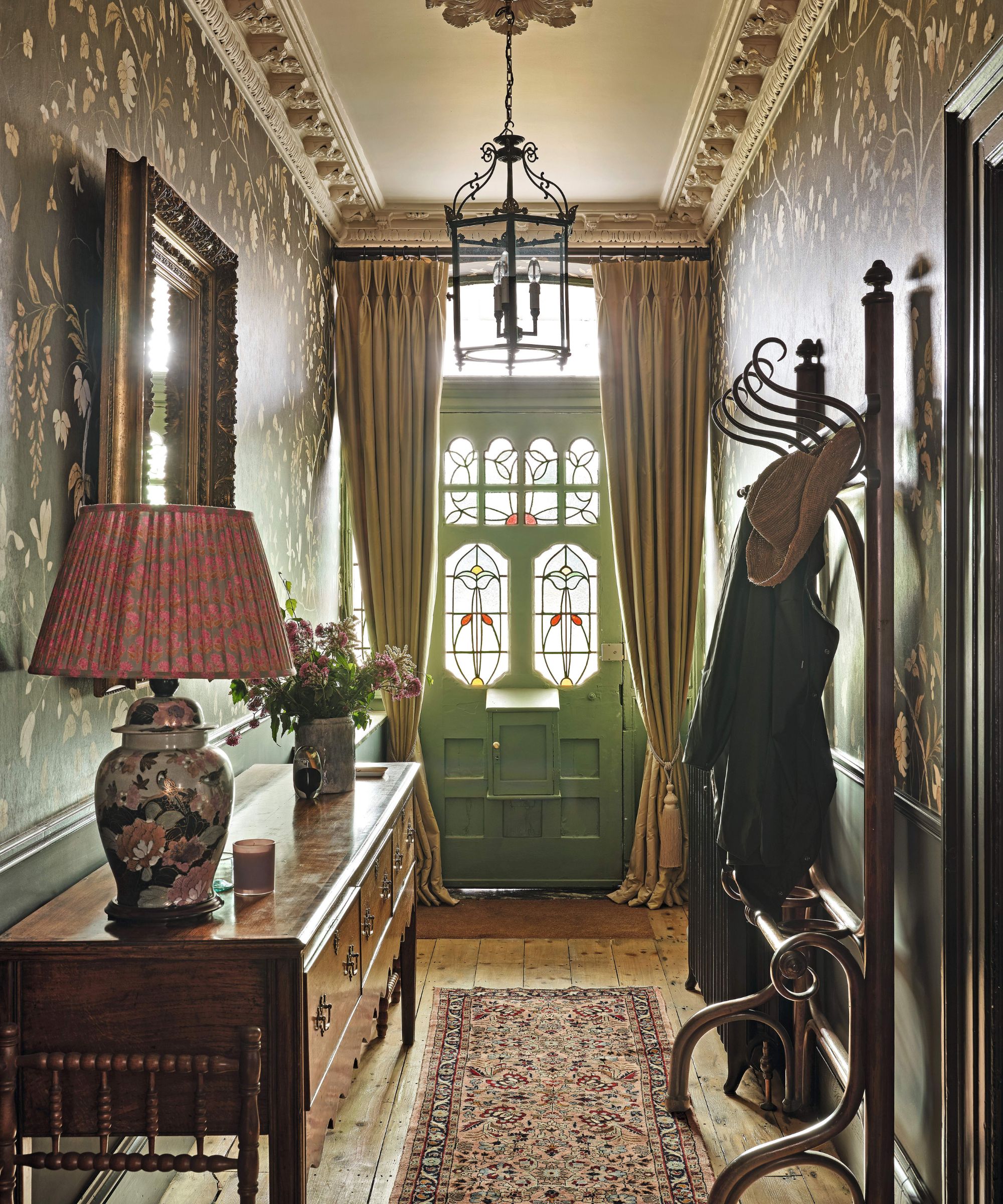 a sunlit wallpapered entryway with traditional Victorian front door, ceiling moldings and paneleding with an antique wooden hall table, rug and coat hooks