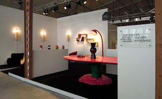 View of Europe's La Galerie Nationale exhibition stand featuring white walls, black flooring, a green and red table with a lamp and a black sculpture on the table