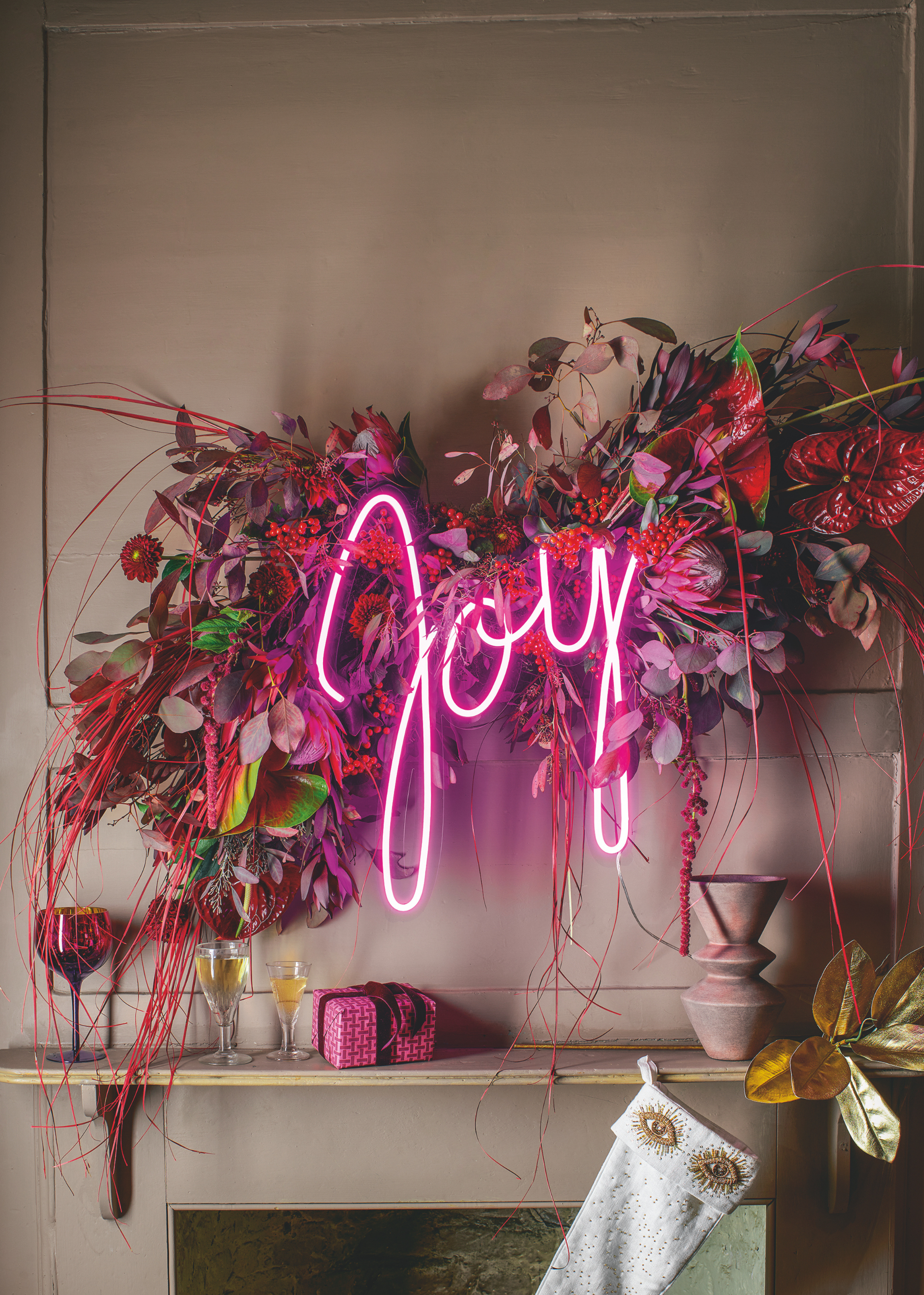 A pink neon 'Joy' sign is lit against a vibrantly colored hanging foliage display above a mantelpiece with a white stocking hanging underneath with gold eyes