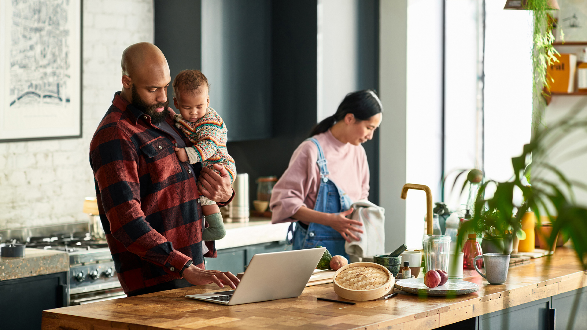 A family in the kitchen: the dad holds the baby as he checks something on his laptop while the mom does the dishes