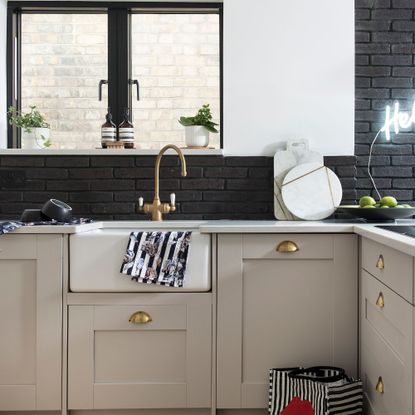 Butler sink in a beige shaker style kitchen with brass accents and a black brick wall