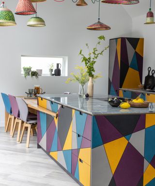 A kitchen island with a yellow, purple, blue, and gray base, a granite worktop, a wooden breakfast bar next to it with purple and blue chairs, and red and green hanging pendant lights above it