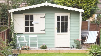 Light green shed at the bottom of a garden with chairs in front of it