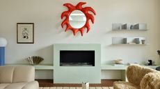 Image of an off-white living room wall with a modern, light mint green fireplace. There is a sculptural, red mirror over the fireplace, a fuzzy accent chair, and chrome shelving in the room.