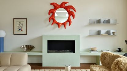 Image of an off-white living room wall with a modern, light mint green fireplace. There is a sculptural, red mirror over the fireplace, a fuzzy accent chair, and chrome shelving in the room.